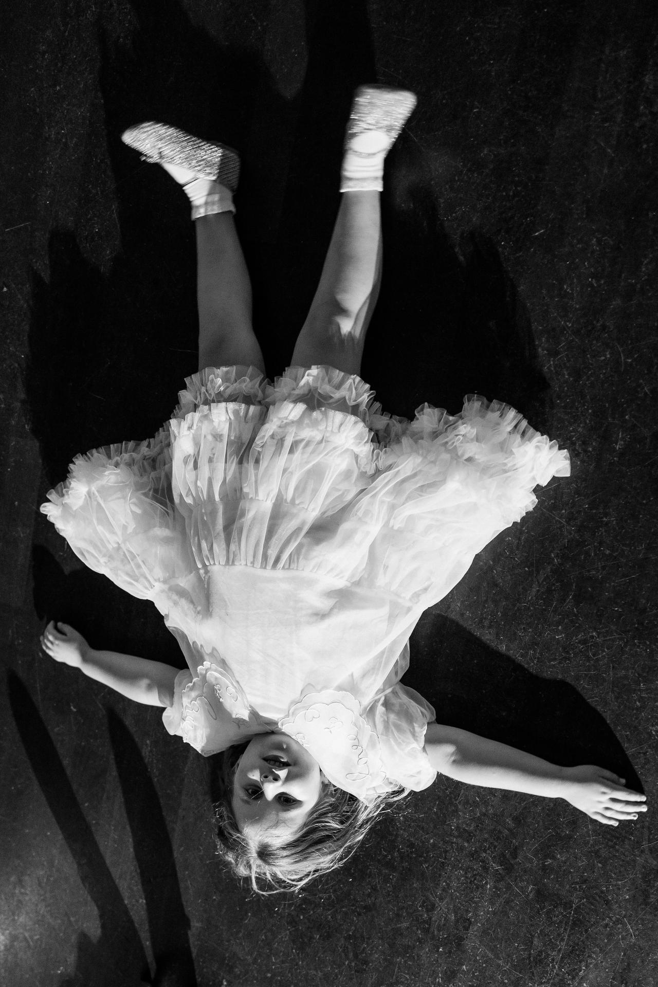 An overhead black-and-white portrait of a flower girl in a white dress, with her arms extended. Wedding photography at Ateres Charna, New Square, NY.