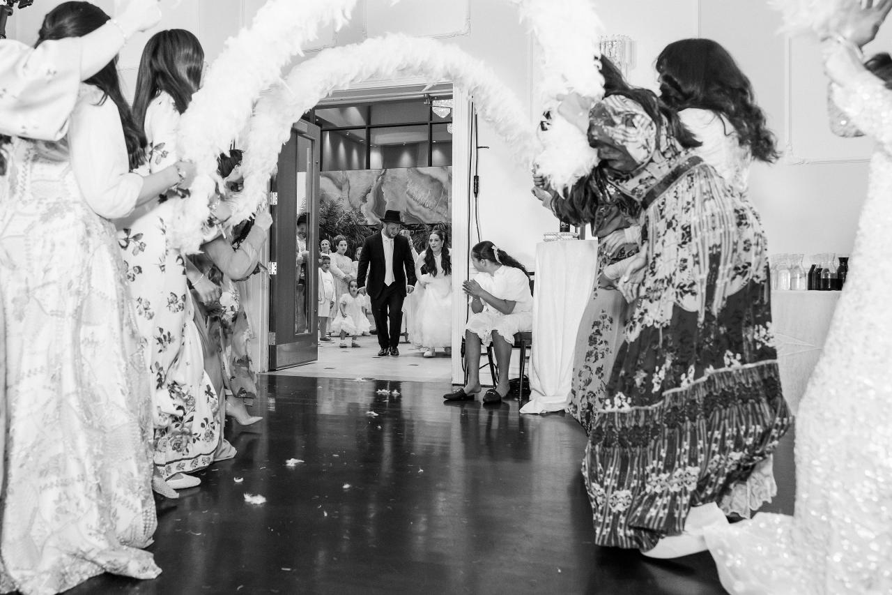 The newlyweds entering the reception through a white floral arch, framed by guests in black and white. Wedding photography at Ateres Charna, New Square, NY.
