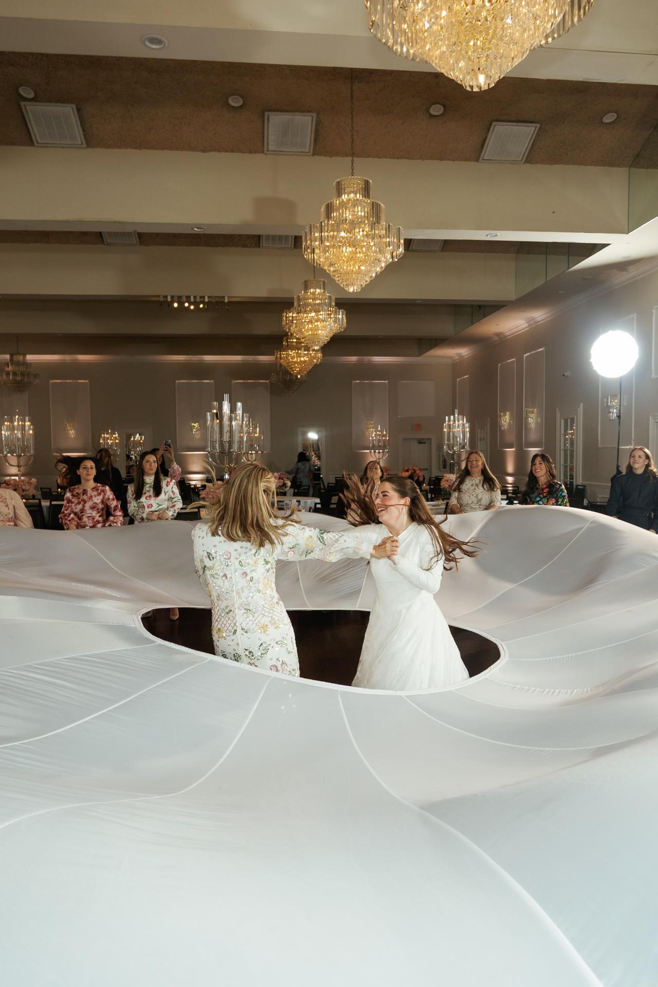A billowing white parachute around the couple and family, with a circular opening and bright folds. Wedding photography at Ateres Charna, New Square, NY.