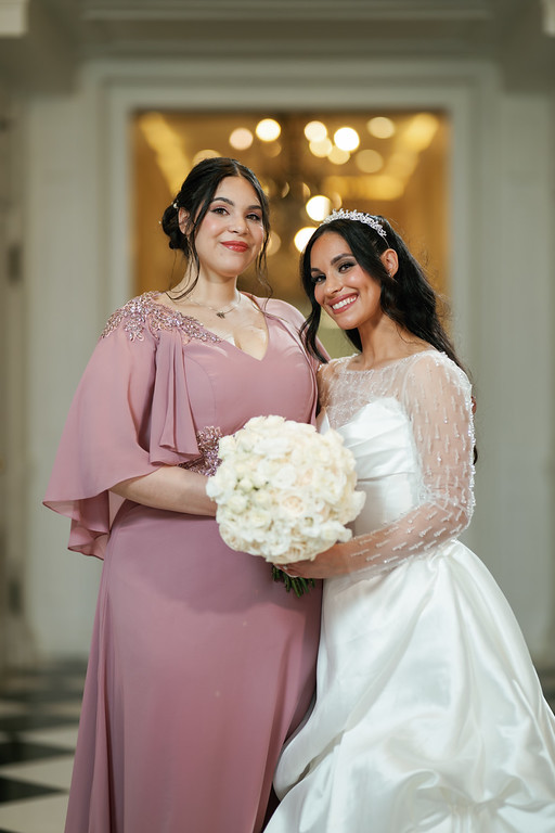 Two women smiling together at a wedding, one in a mauve dress and the other in a white dress holding a bouquet in a softly lit venue hallway.
