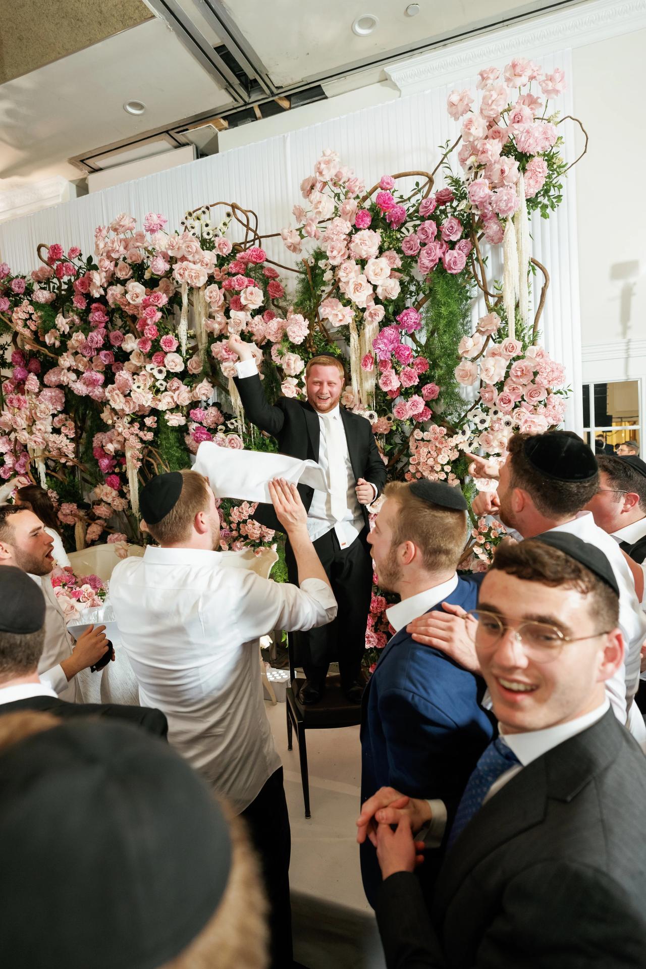 A wide wedding celebration against the flower wall, with raised arms and layers of guests. Wedding photography at Ateres Charna, New Square, NY.