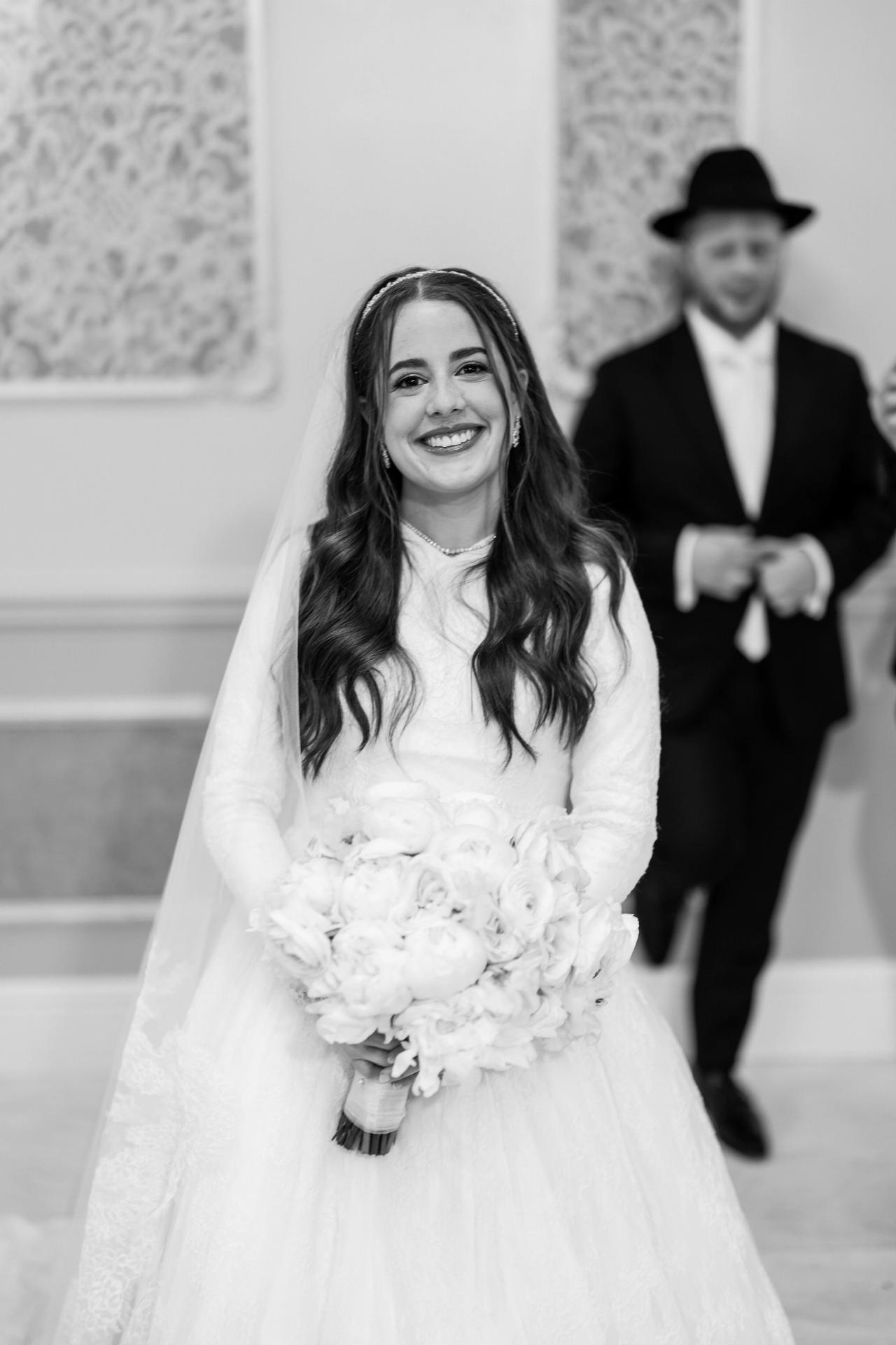 A black-and-white bridal portrait on a staircase, with the groom softly visible above. Wedding photography at Ateres Charna, New Square, NY.