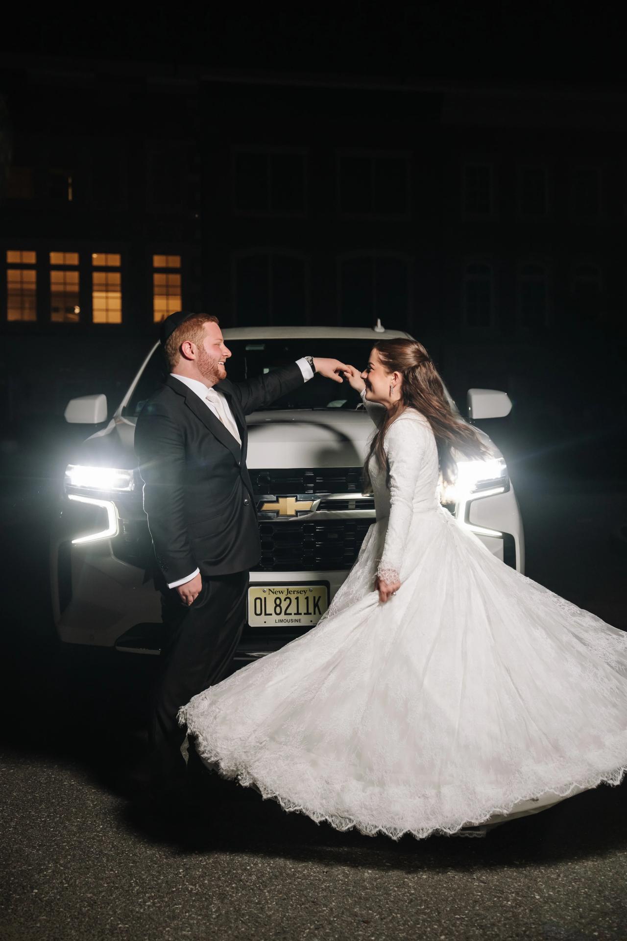 The bride twirling beside a car in headlight glow at night, from the Ateres Charna wedding portrait collection. Wedding photography.