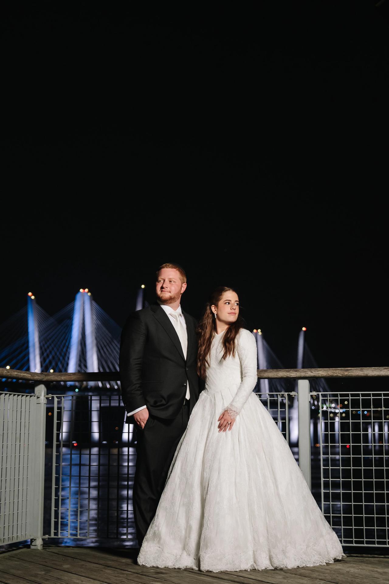 The newlyweds beside water with illuminated bridge cables behind them at night, from the Ateres Charna wedding portrait collection. Wedding photography.