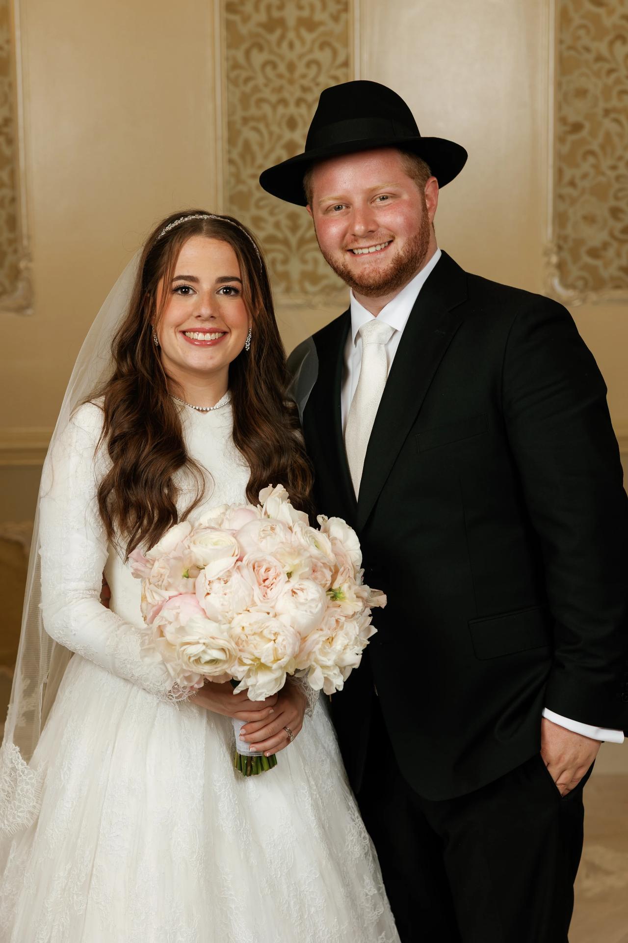 A warm close portrait of the smiling newlyweds, with the bouquet in the foreground. Wedding photography at Ateres Charna, New Square, NY.