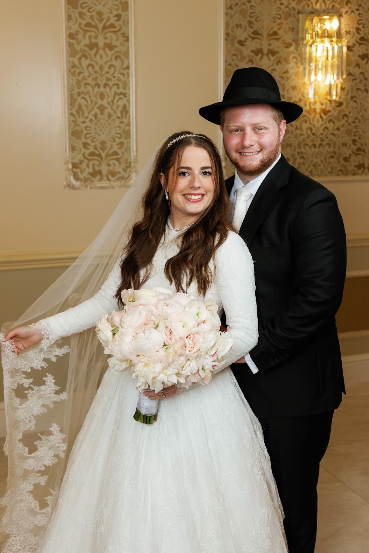 A warm indoor portrait of the newlyweds with lace veil and bouquet against ornate gold wall panels. Wedding photography at Ateres Charna, New Square, NY.