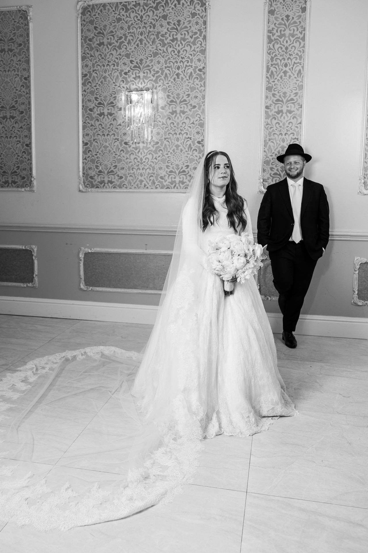 A balanced black-and-white portrait of the newlyweds, with the wedding gown and interior architectural lines leading through the frame. Wedding photography at Ateres Charna, New Square, NY.