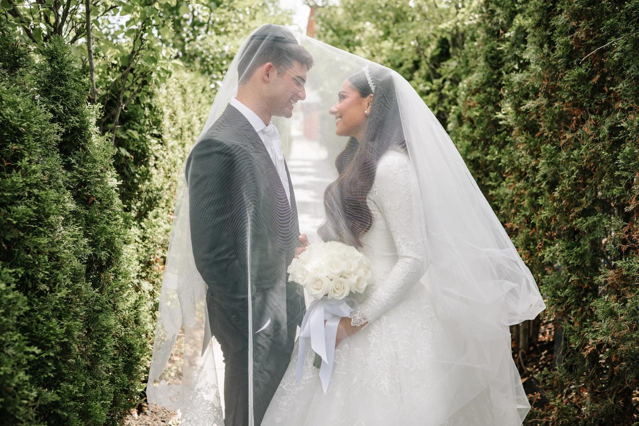 The couple shares a veil in a hedge-lined passage. Wedding photography at Brightstone, Passaic, NJ.