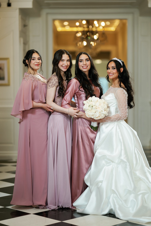 A group of four women pose for a formal portrait in a ballroom hallway; the bride in a white gown holds a bouquet while the others wear coordinated mauve dresses.