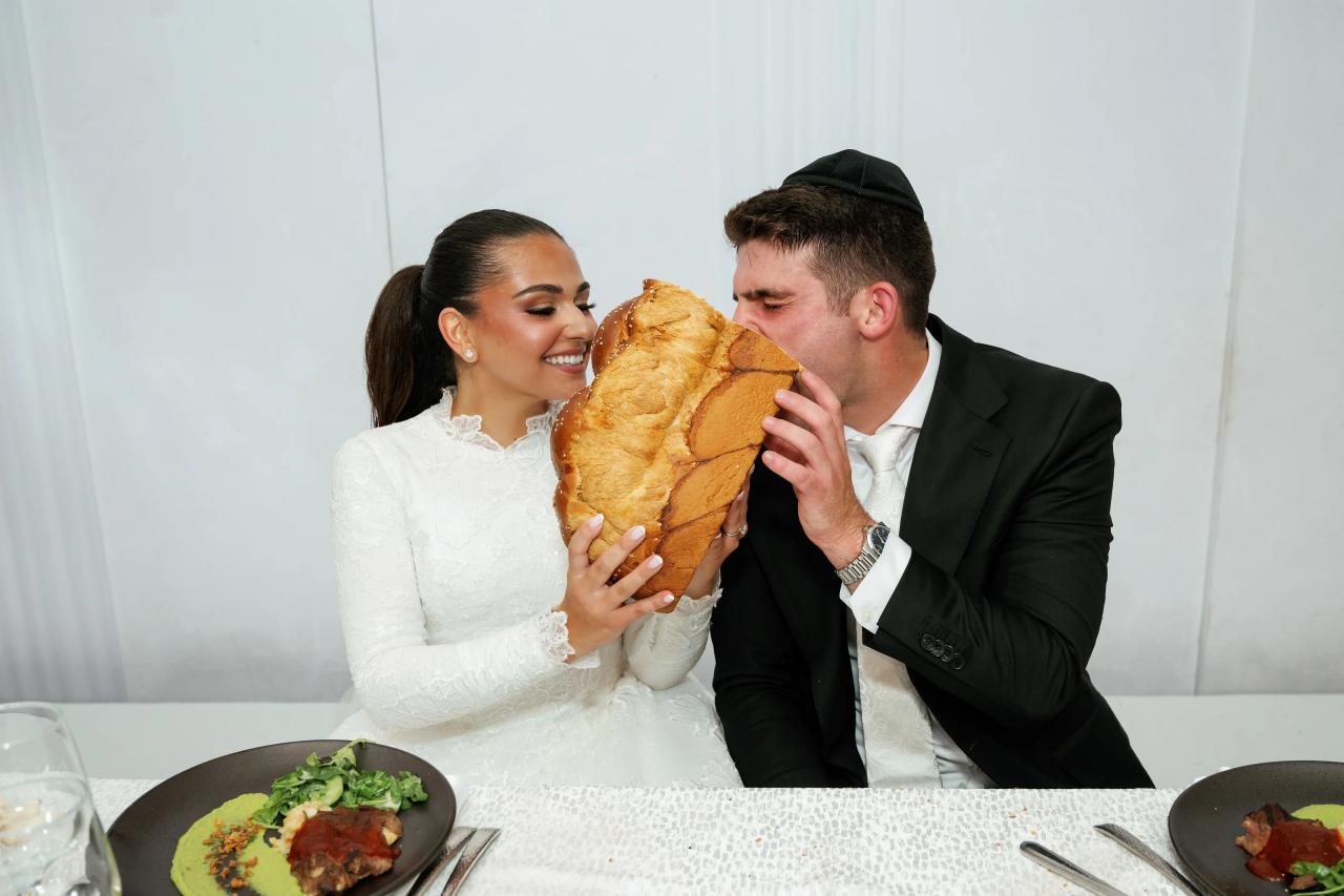 The newlyweds share a playful challah moment at the reception table. Wedding photography at Brightstone, Passaic, NJ.