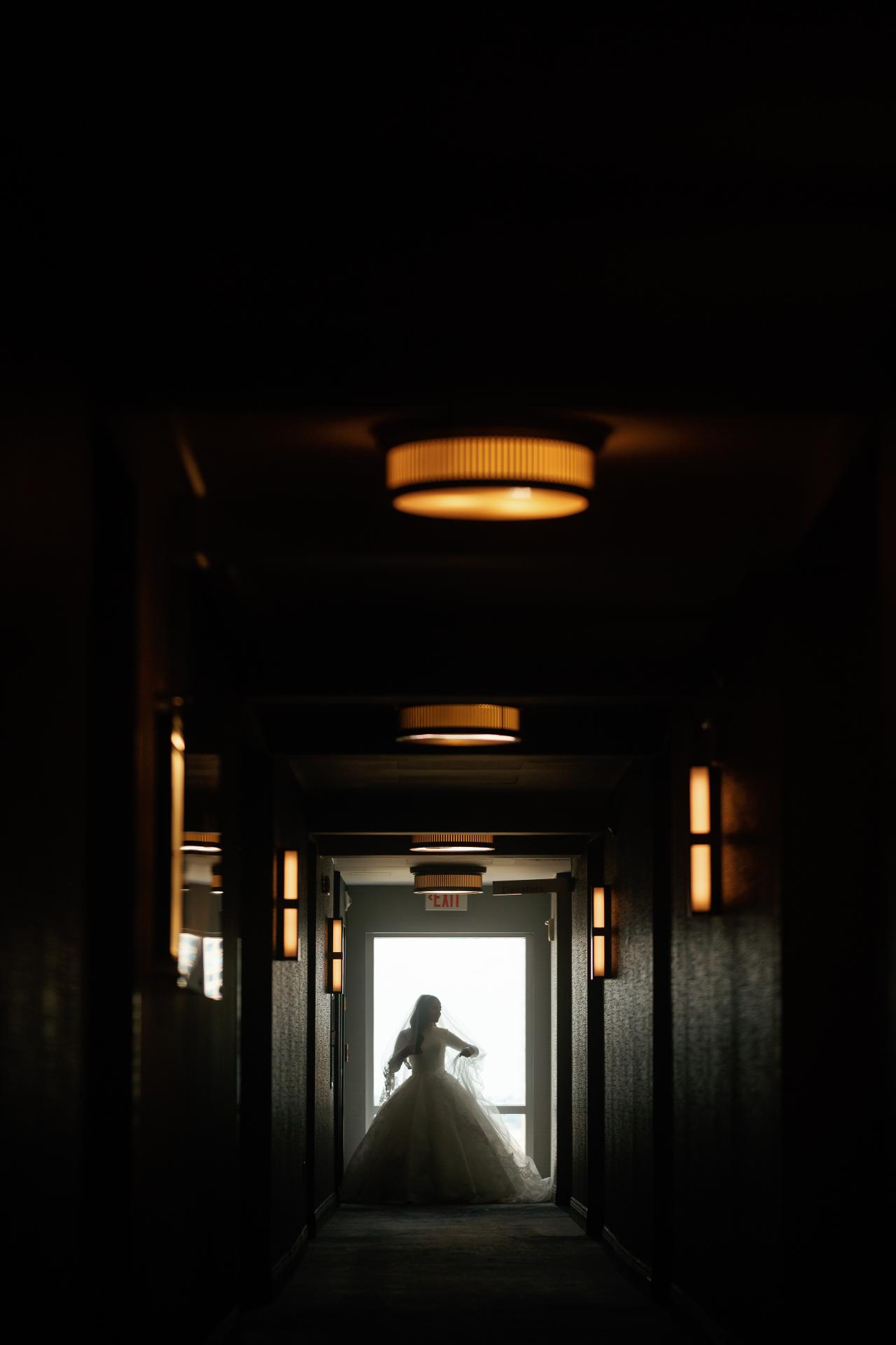 Backlit bride silhouette at the end of a dark corridor, framed by repeating ceiling fixtures. Wedding photography at Hilton Baltimore Inner Harbor, Baltimore, MD.