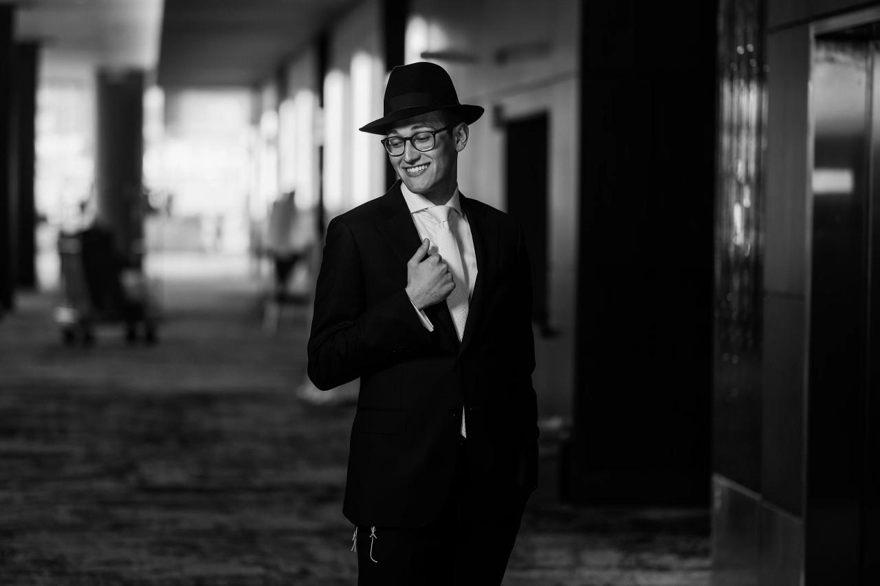 Relaxed black-and-white groom portrait with receding hotel corridor lights. Wedding photography at Hilton Baltimore Inner Harbor, Baltimore, MD.
