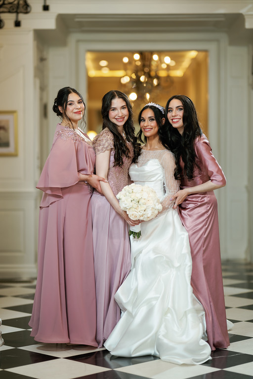 Four women pose for a formal wedding portrait with the bride in the center holding a white bouquet, indoors in front of a chandelier-lit entryway.