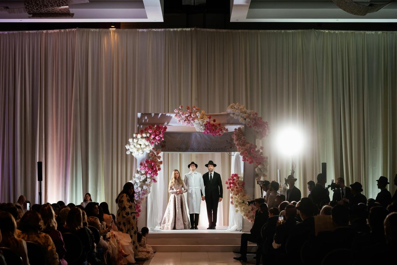 Wide floral chuppah scene across the guests, with dramatic light against tall draped walls. Wedding photography at Hilton Baltimore Inner Harbor, Baltimore, MD.