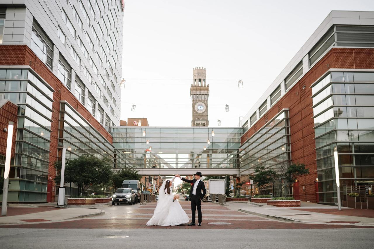 Wide architectural couple portrait between Hilton buildings, with the Baltimore clock tower and glass skybridge above — couple portraits for the Hilton Baltimore Inner Harbor wedding. Wedding photography.