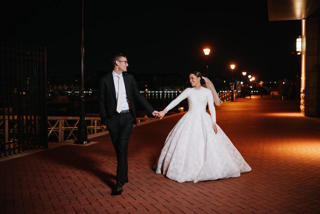 Couple walking hand in hand by the waterfront after dark, warm streetlights receding behind them — couple portraits for the Hilton Baltimore Inner Harbor wedding. Wedding photography.