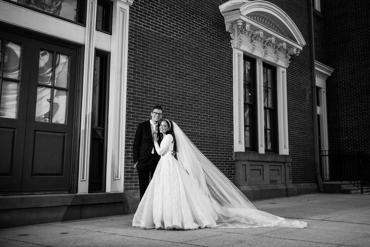 Wide black-and-white couple portrait at a historic brick facade, with veil trailing across the pavement — couple portraits for the Hilton Baltimore Inner Harbor wedding. Wedding photography.