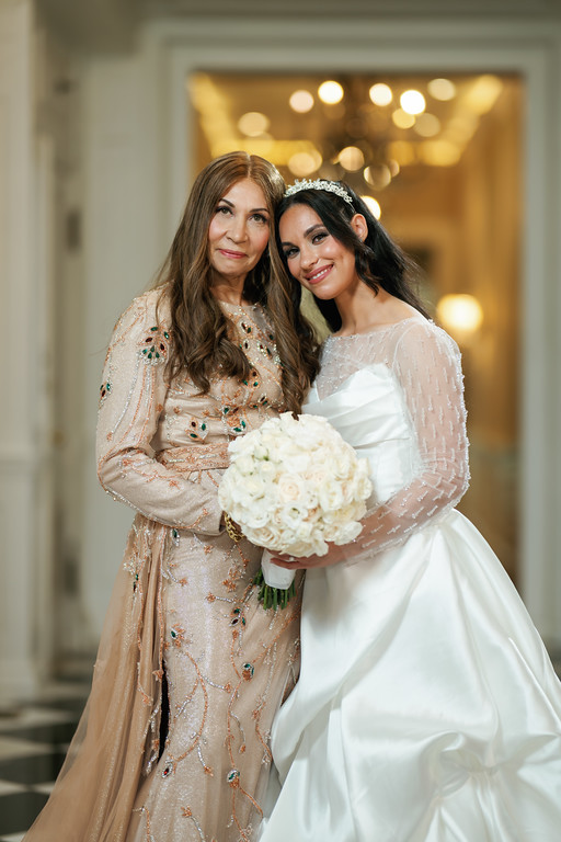 Smiling bride in a white gown holding a bouquet embraces an older woman in a beaded dress in a warmly lit venue hallway.