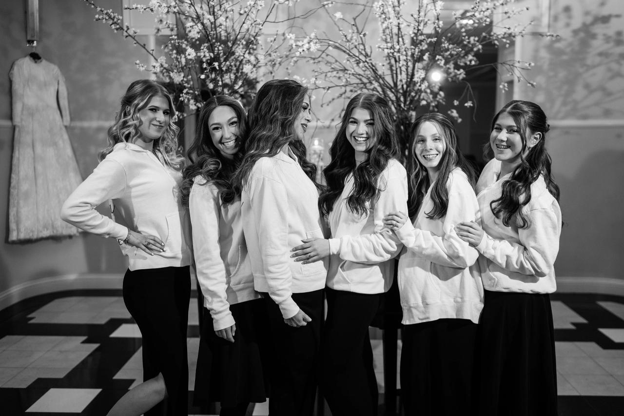 Black-and-white photo of six women smiling and laughing together in matching outfits outdoors at a simcha, standing close as if posing mid-celebration. Wedding photography at The Palace, Brooklyn, NY.