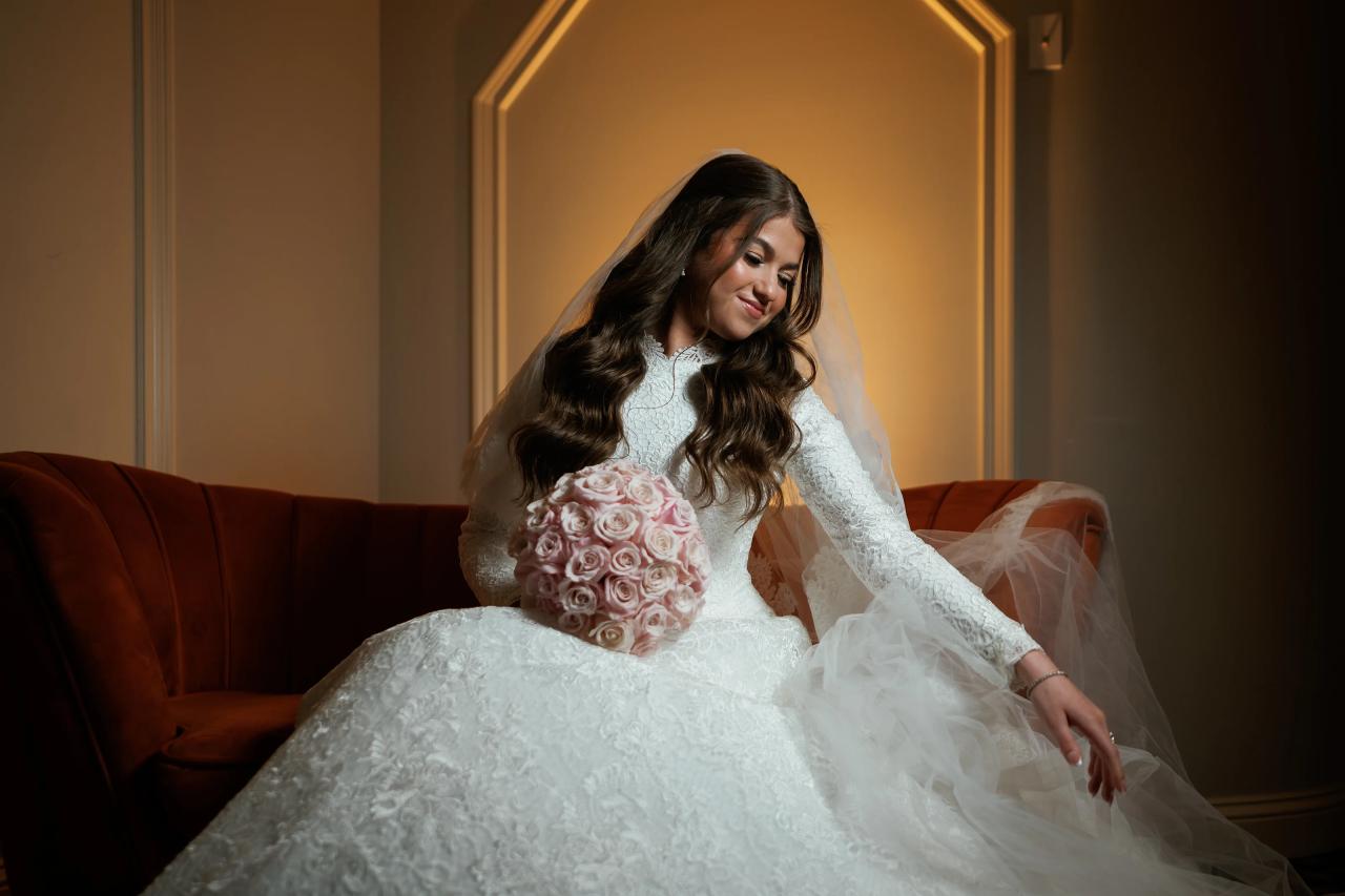 Bride seated on a tufted lounge sofa, holding a pink rose bouquet, with a soft smile and veil draped over her shoulders under warm amber light. Wedding photography at The Palace, Brooklyn, NY.