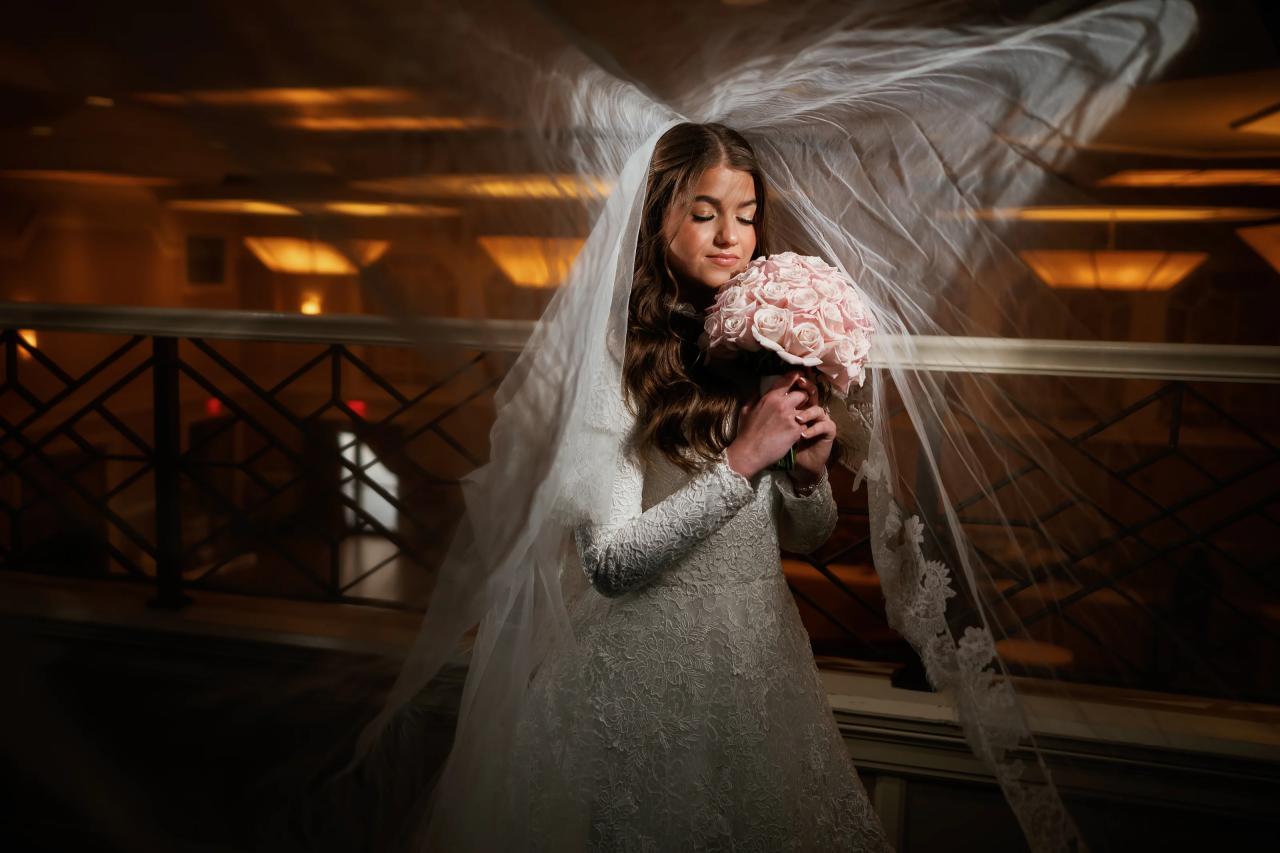 Bride in a lace gown holding a pink rose bouquet under warm amber light, veil billowing as she stands by an upper balcony railing. Wedding photography at The Palace, Brooklyn, NY.