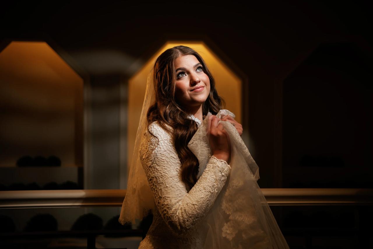 Bride in lace wedding gown adjusting her veil under warm amber architectural light, looking up with a soft smile. Wedding photography at The Palace, Brooklyn, NY.