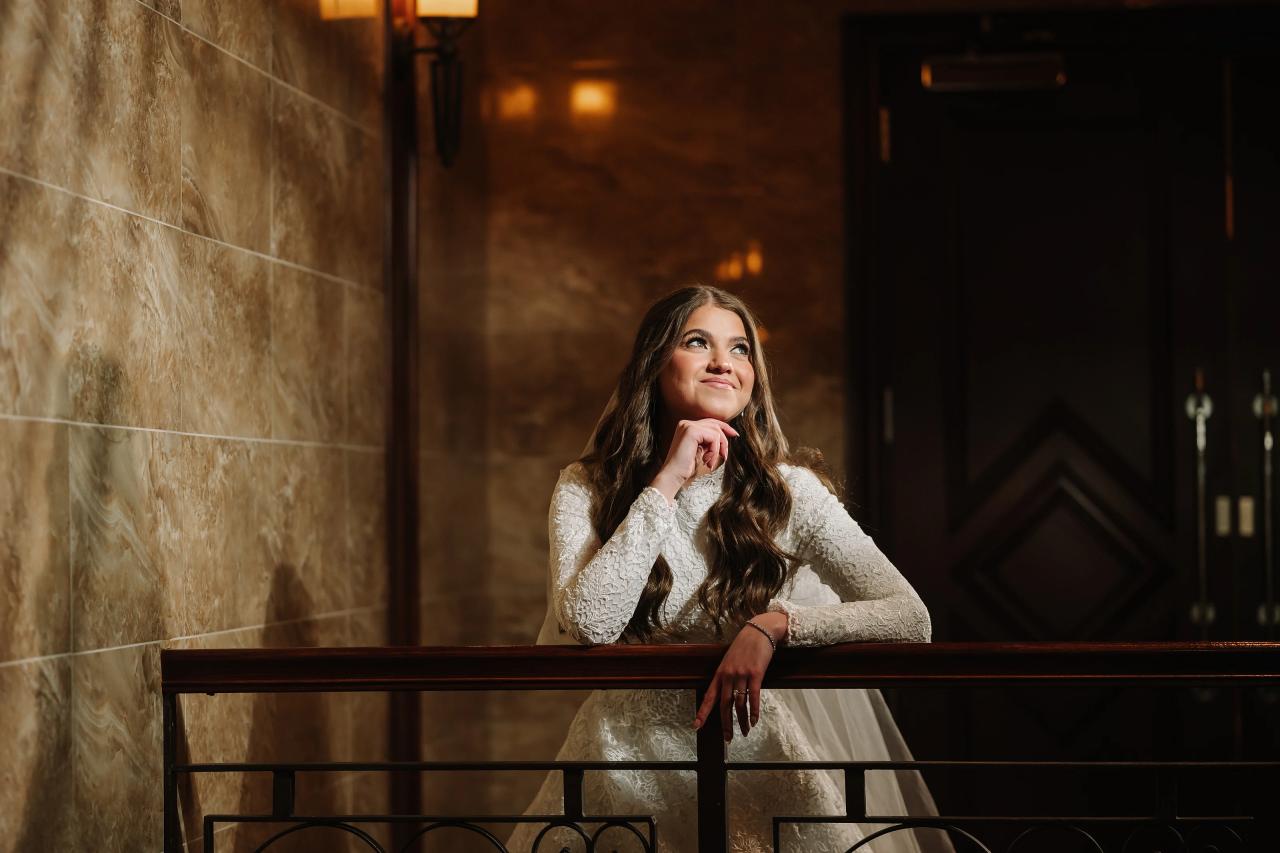 Bride in lace leaning on an ornate railing in a warm, dimly lit interior, gazing upward with a calm expression. Wedding photography at The Palace, Brooklyn, NY.