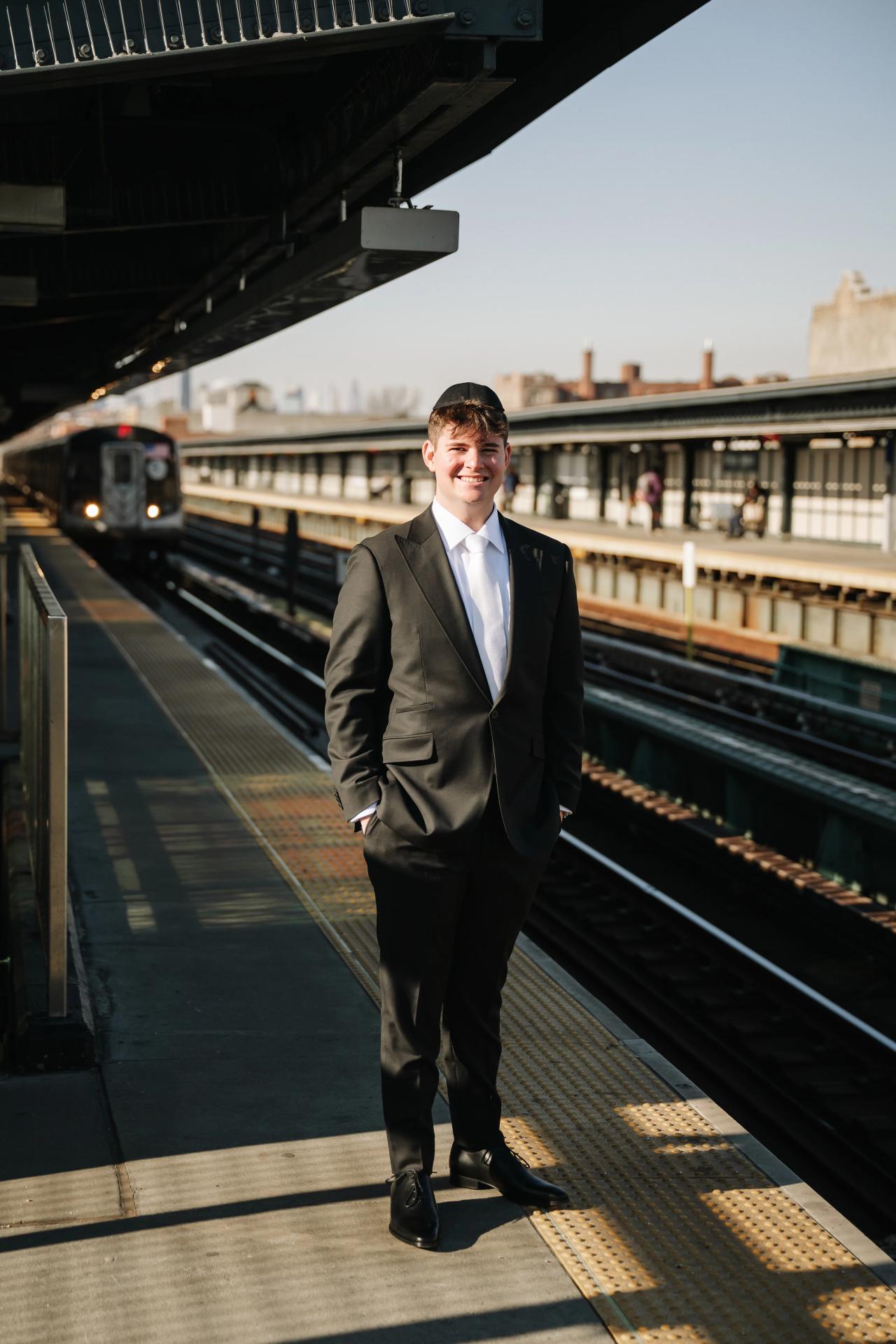 Smiling groom in a dark suit standing on a train platform with tracks and canopy lines receding behind him. Wedding photography at The Palace, Brooklyn, NY.