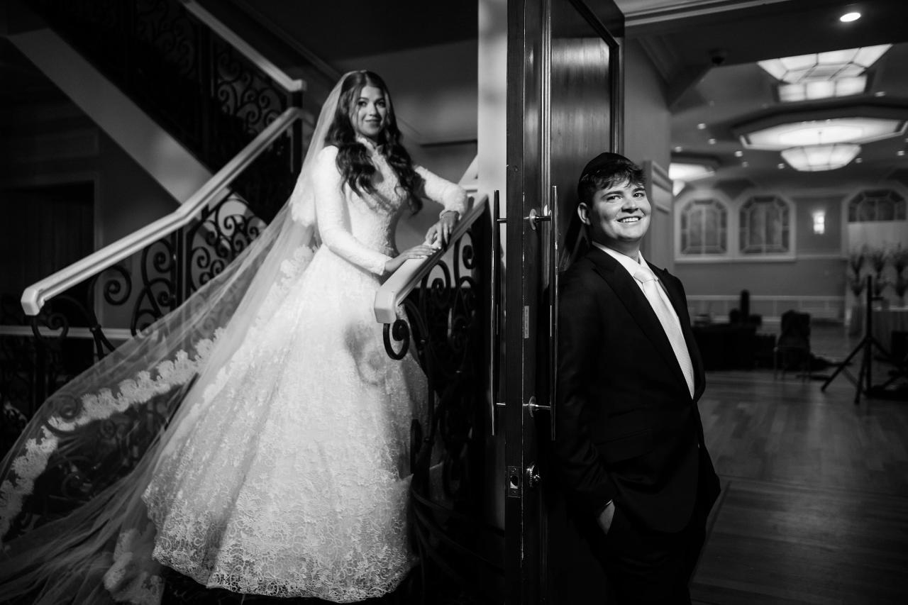 Black-and-white candid of a bride in a lace gown and veil stepping through a doorway with the groom in a dark suit, both smiling in a quiet hallway moment. Wedding photography at The Palace, Brooklyn, NY.