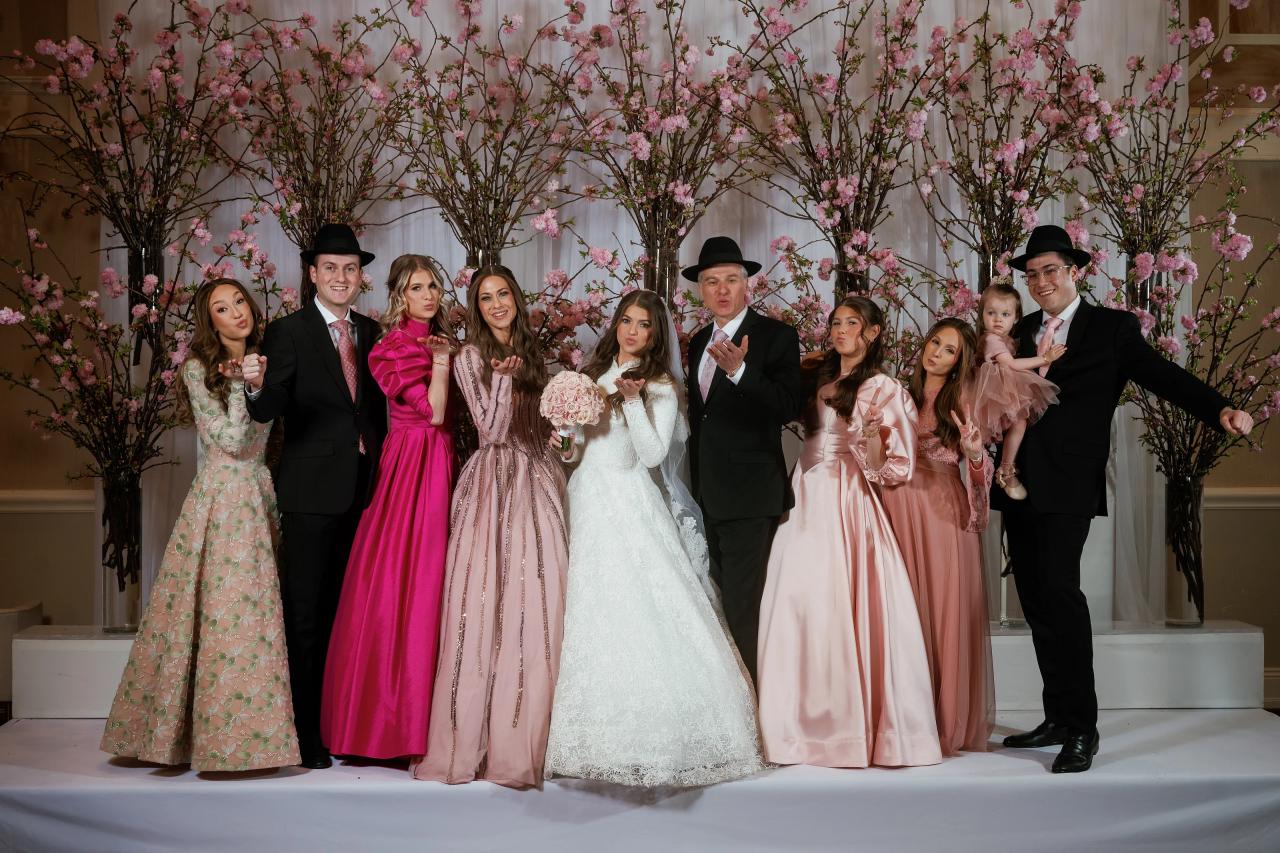 Group of family and friends in formal clothing posing on a stage with a pink floral backdrop, with the bride in white centered in the group and hands raised toward the camera. Wedding photography at The Palace, Brooklyn, NY.