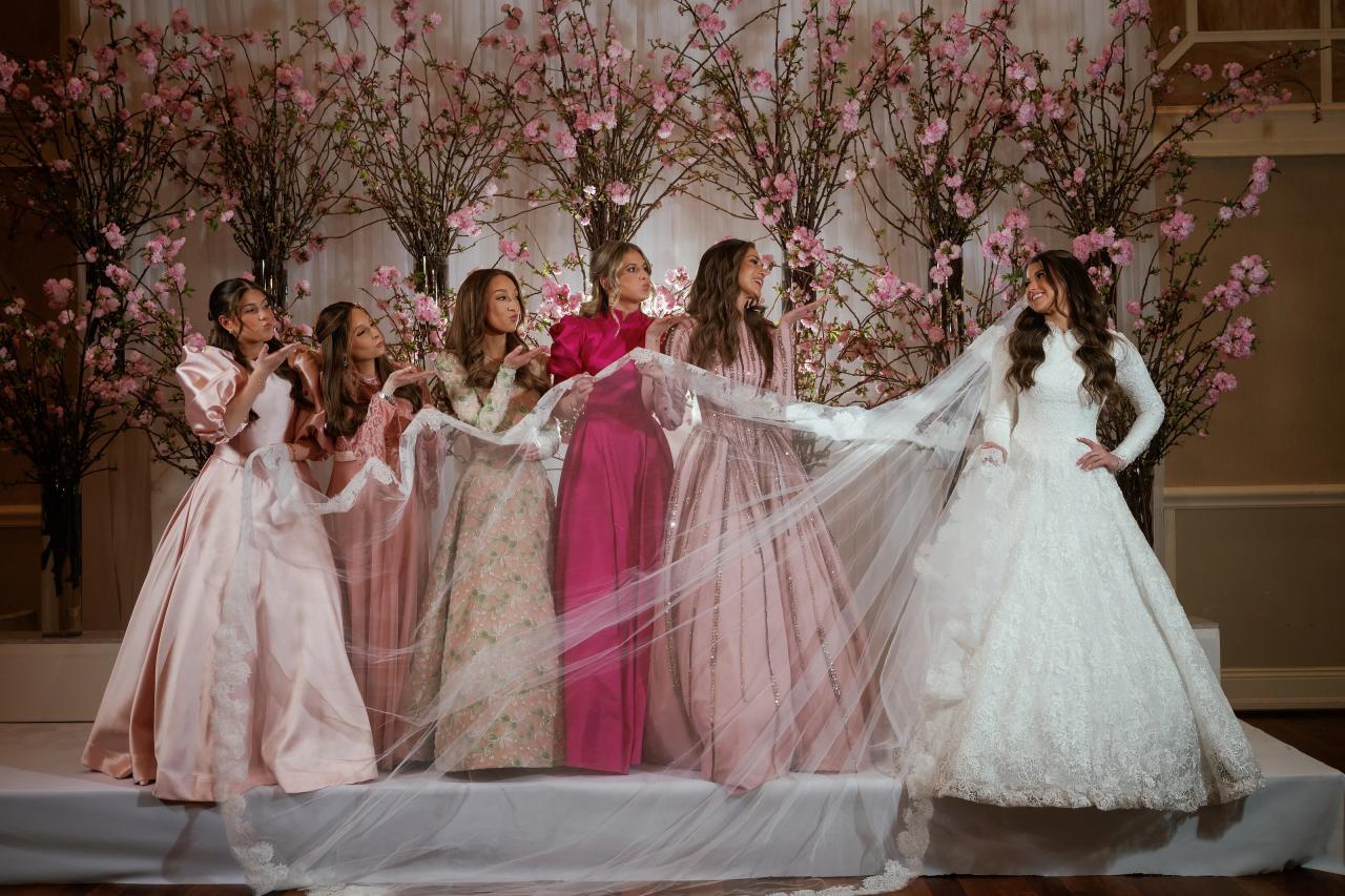 Bride in a white wedding dress twirling on a stage with a flowing veil, surrounded by friends in pink and pastel dresses; pink blossom décor behind them. Wedding photography at The Palace, Brooklyn, NY.