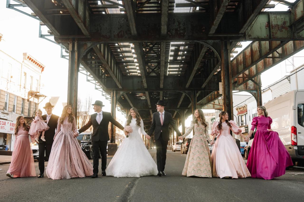 Wedding couple and wedding party walking toward the camera under an industrial steel walkway, with bridesmaids in pastel pink and floral dresses and soft daylight. Wedding photography at The Palace, Brooklyn, NY.