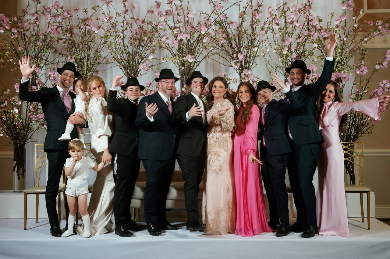 Group of guests on an event stage with pink floral backdrop, many smiling with hands raised, including a bride and groom among the group. Wedding photography at The Palace, Brooklyn, NY.