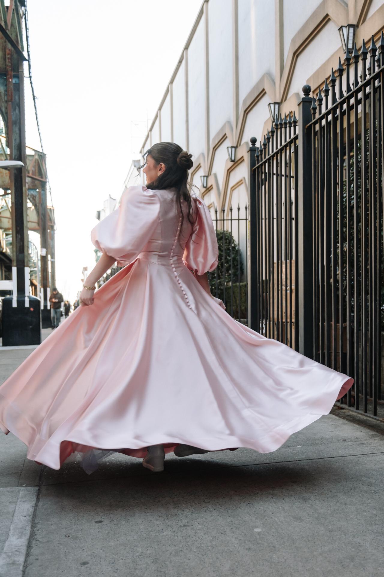 Bride in a light pink dress twirls outdoors, seen from behind, her flowing skirt and veil catching the light against a black iron fence. Wedding photography at The Palace, Brooklyn, NY.
