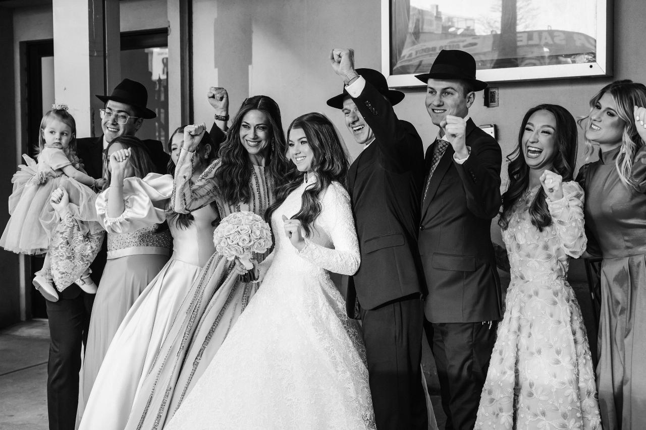 Black-and-white dance-floor moment: bride holding her bouquet smiling toward the camera while the groom and guests cheer around her with raised hands. Wedding photography at The Palace, Brooklyn, NY.