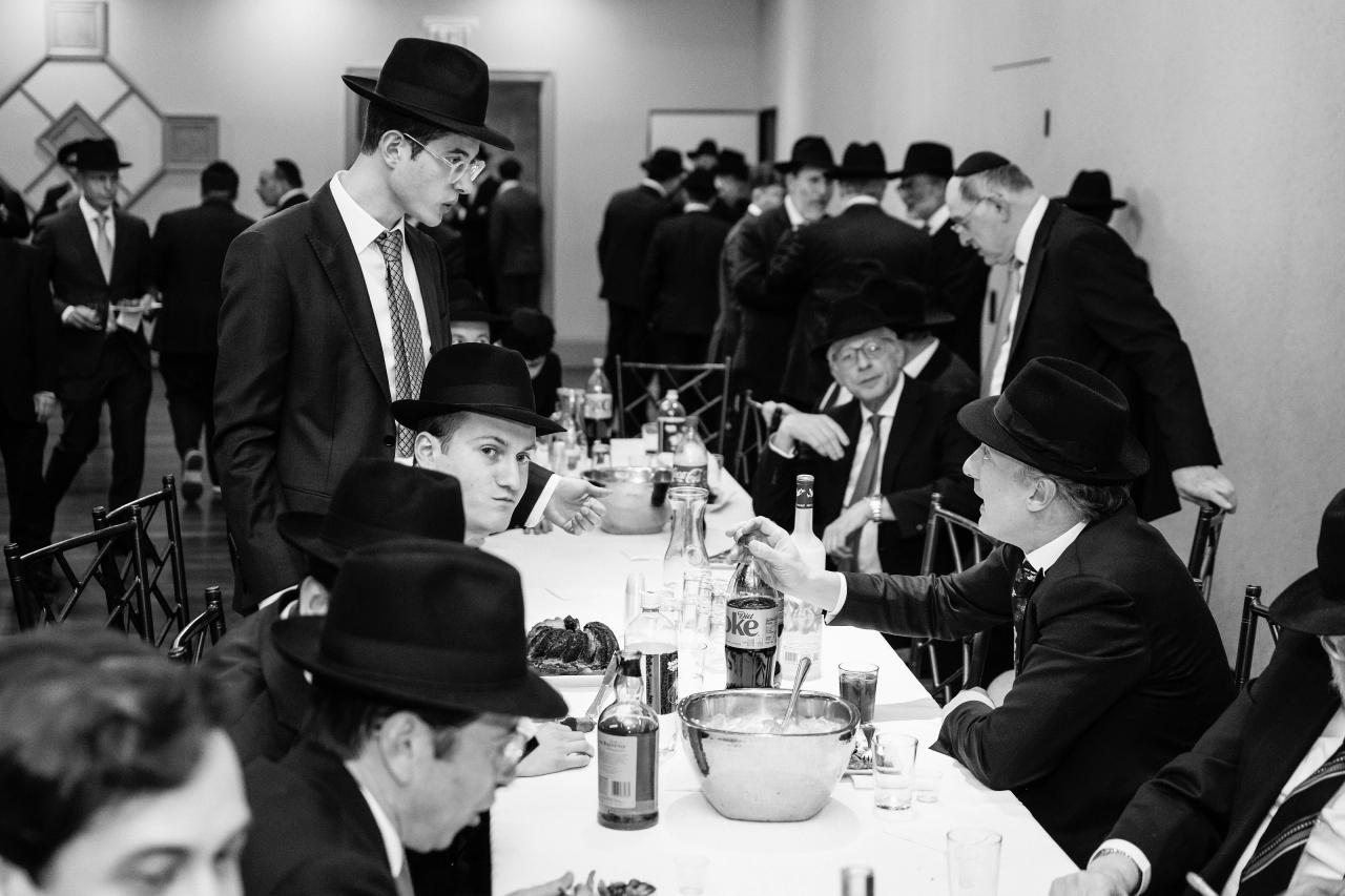 Black-and-white scene of guests in hats seated at a reception table, bottles and glasses in the foreground, one man reaching across mid-conversation. Wedding photography at The Palace, Brooklyn, NY.