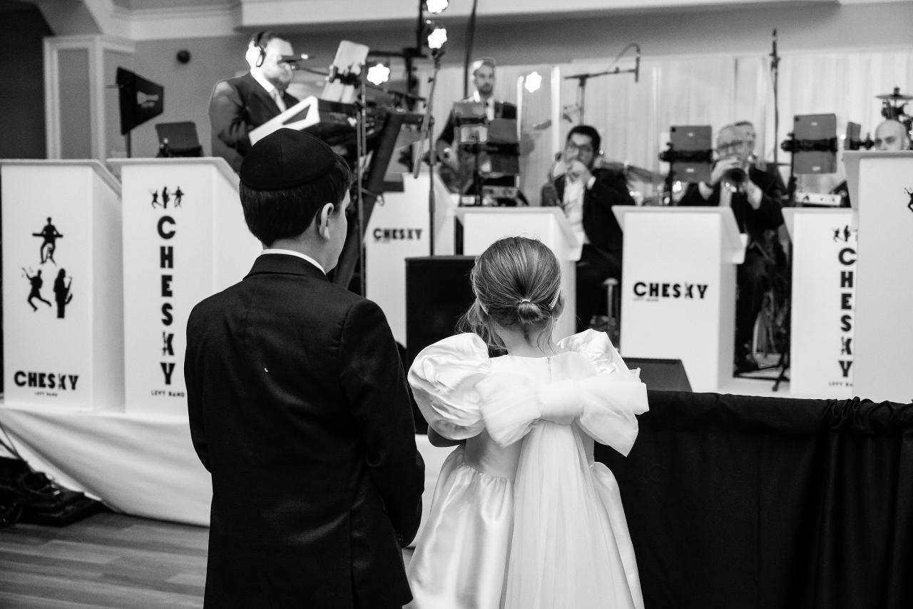 Black-and-white candid of a bride and groom from behind standing close and facing a live band during their celebration, with the reception stage in view. Wedding photography at The Palace, Brooklyn, NY.