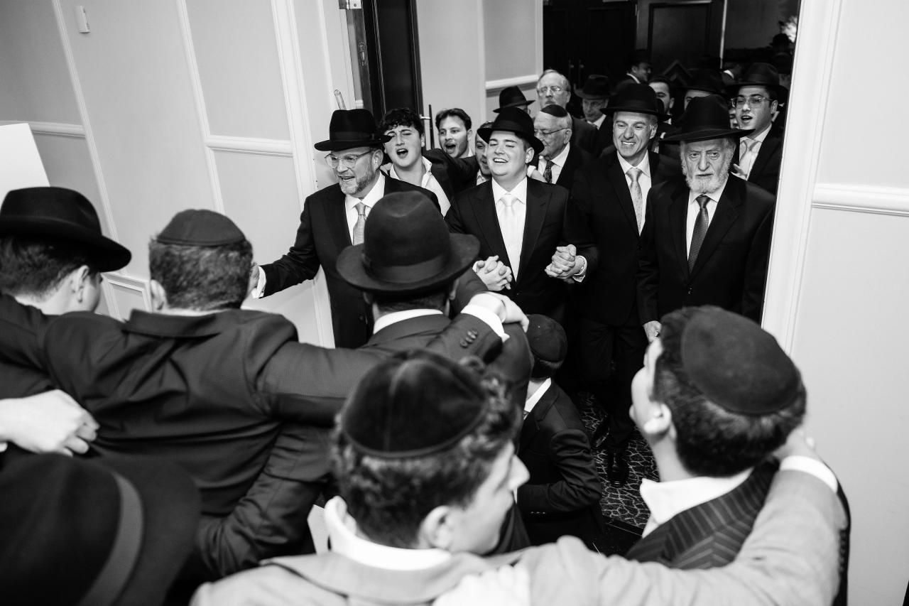Black-and-white candid of a wedding celebration crowd, with the couple near the center laughing and reaching through the surrounding guests in formalwear and hats. Wedding photography at The Palace, Brooklyn, NY.