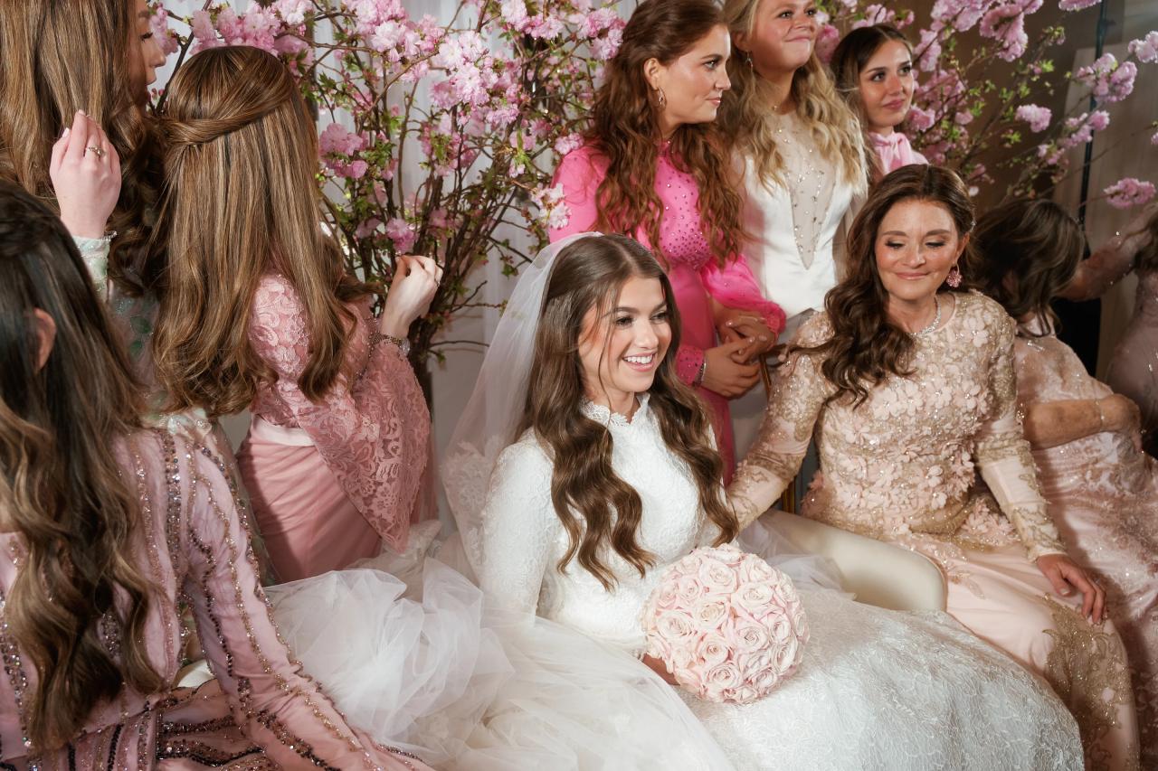 Wedding reception: bride seated in front of a pink floral backdrop surrounded by friends and family, smiling with a bouquet and veil. Wedding photography at The Palace, Brooklyn, NY.