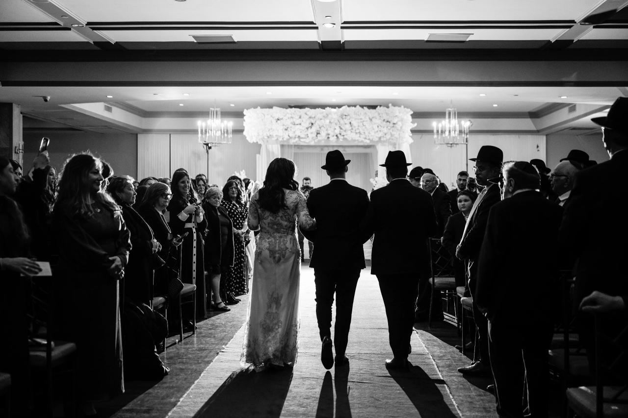 Black-and-white wedding ceremony aisle shot: bride and groom walking forward down the center aisle toward a floral chuppah, guests seated on both sides. Wedding photography at The Palace, Brooklyn, NY.