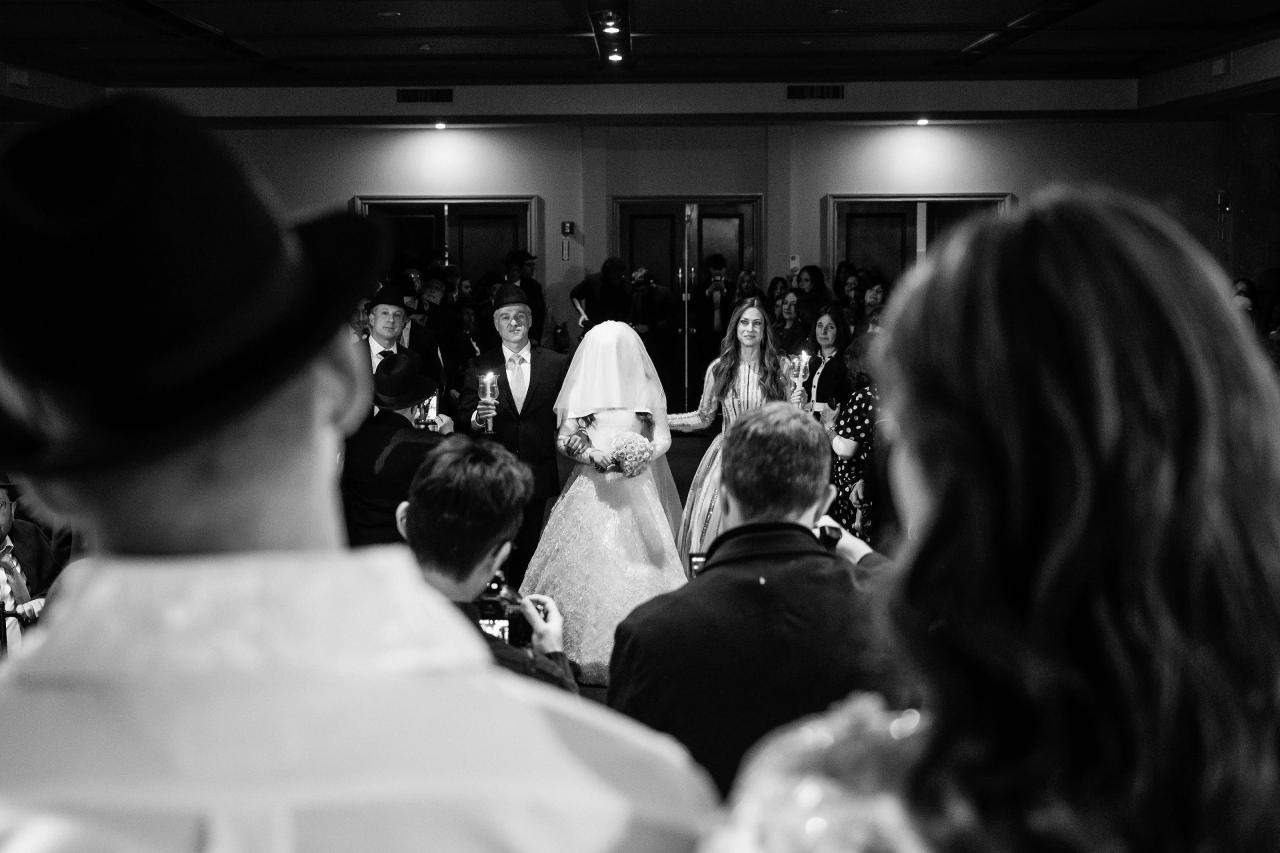 Black-and-white ceremony moment: bride in a white veil and groom in dark attire framed by guests, with audience silhouettes and lit candles in the foreground. Wedding photography at The Palace, Brooklyn, NY.
