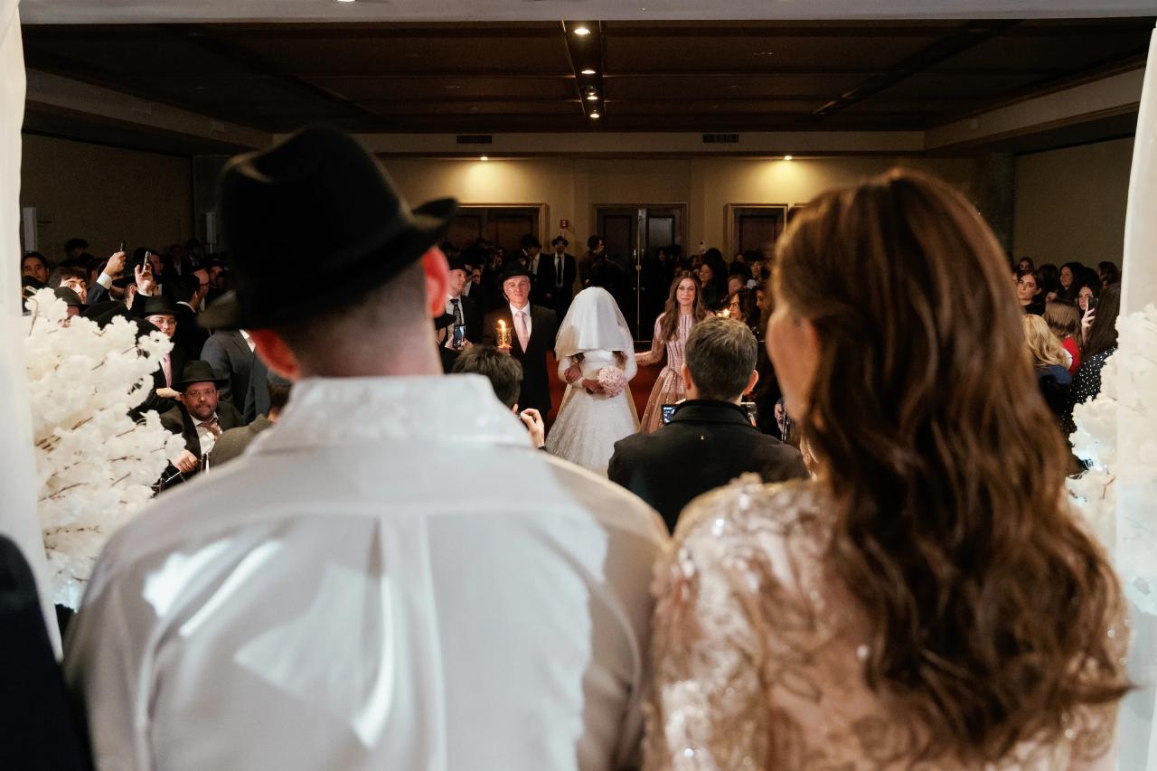 A view from behind guests toward the chuppah, with the bride and groom standing in the center while people in the foreground watch. Wedding photography at The Palace, Brooklyn, NY.