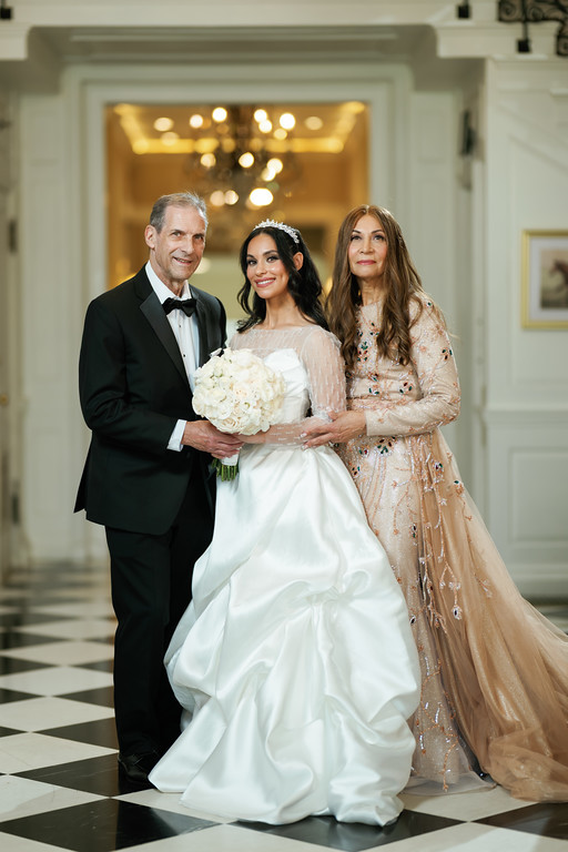 Wedding portrait of a bride in a white gown holding a bouquet, standing between two family members in formal attire in an elegant hallway.