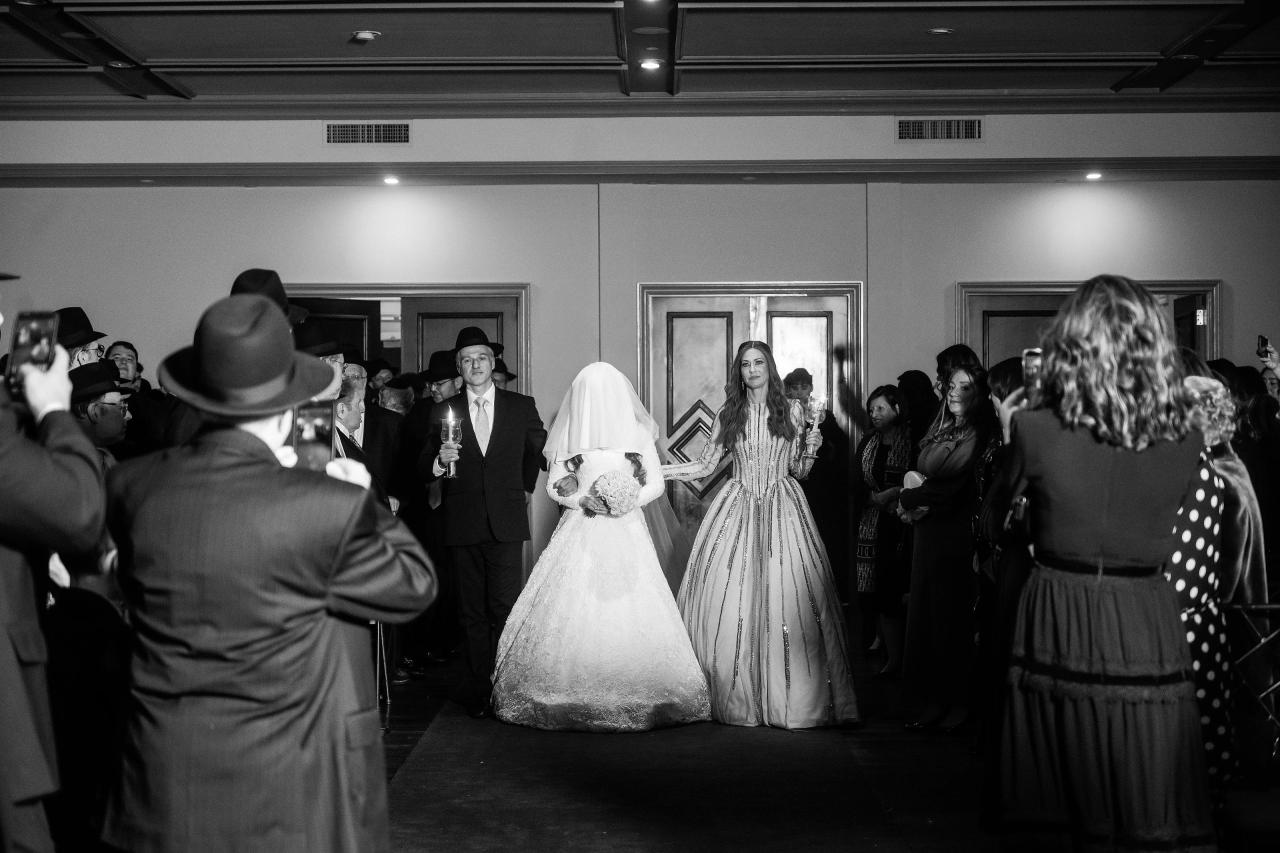 Black-and-white reception moment: bride in a sparkling gown stands centered in a hallway as guests on both sides clap and watch, with raised cameras in the foreground. Wedding photography at The Palace, Brooklyn, NY.