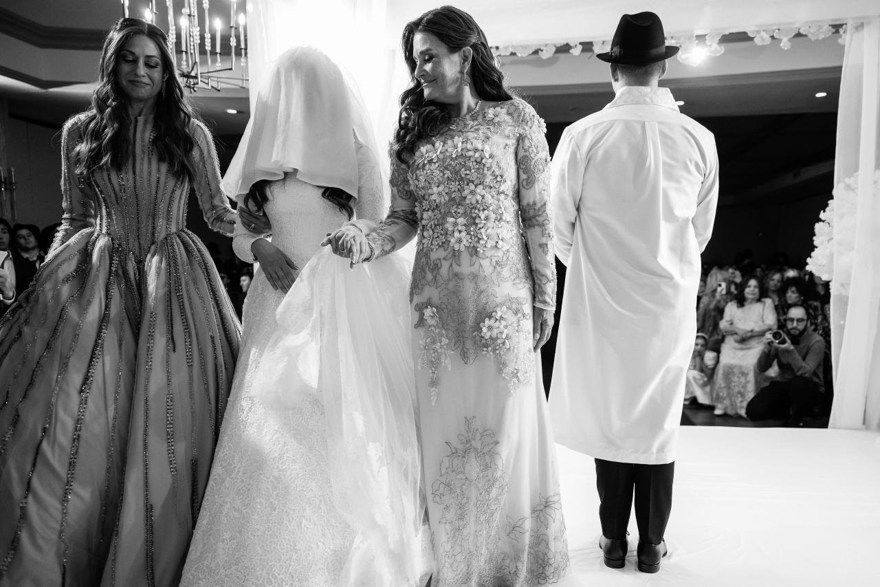 Black-and-white photo of the bride walking forward between family members during a Jewish celebration, with guests and lighting in the background. Wedding photography at The Palace, Brooklyn, NY.