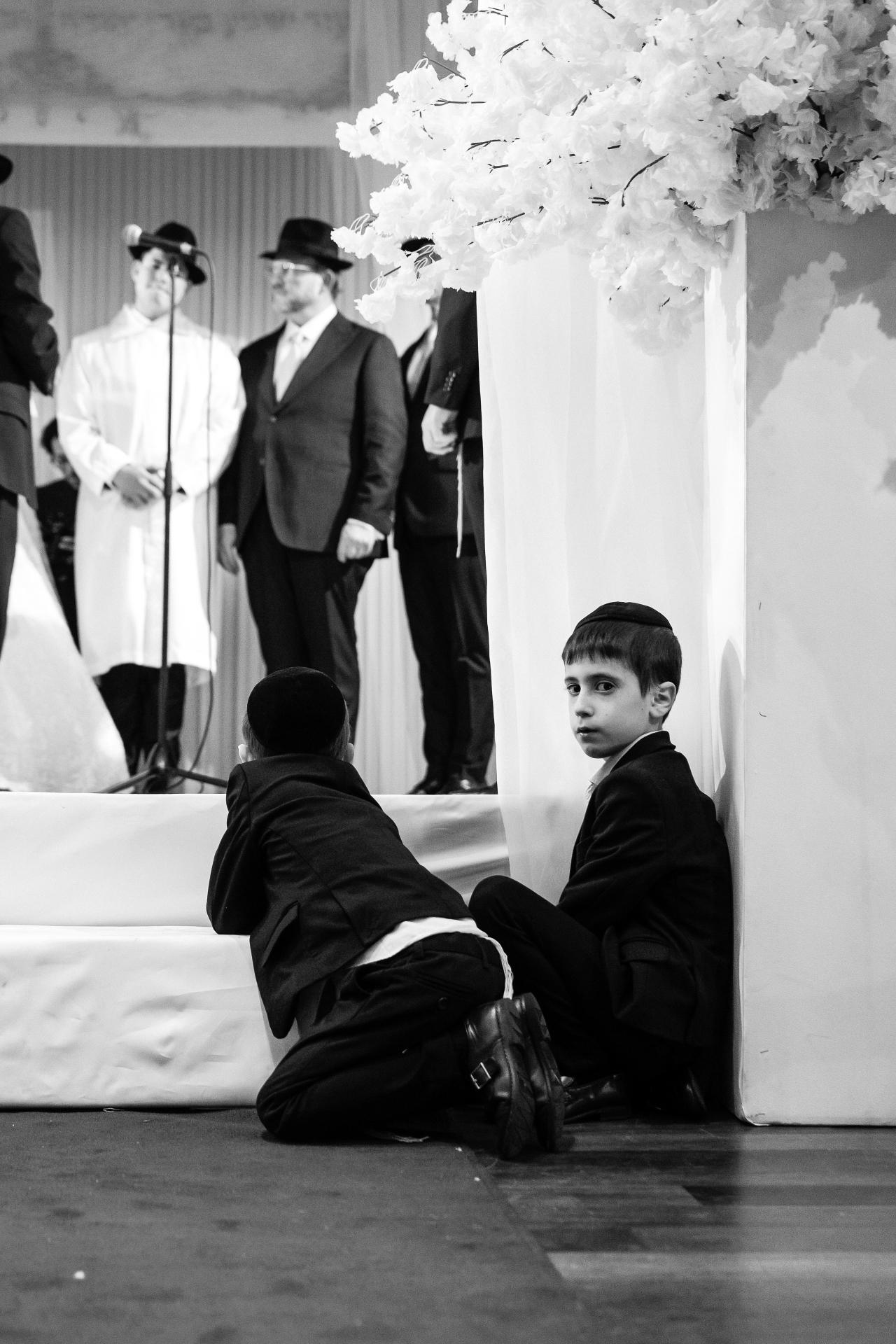Black-and-white wedding moment: a suited young boy sitting on the floor watching the ceremony, with stage guests standing in the background and floral décor above. Wedding photography at The Palace, Brooklyn, NY.