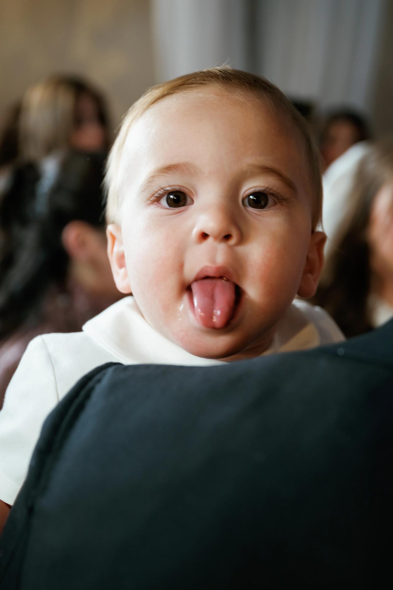 Close-up of a baby making an excited tongue-out face with a blurred crowd in the background. Wedding photography at The Palace, Brooklyn, NY.