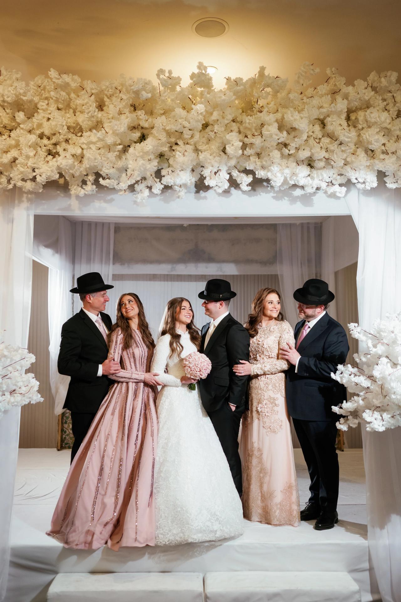 Wedding family and wedding party posed together on a decorated stage with white drapery and cream floral arrangement overhead. Wedding photography at The Palace, Brooklyn, NY.