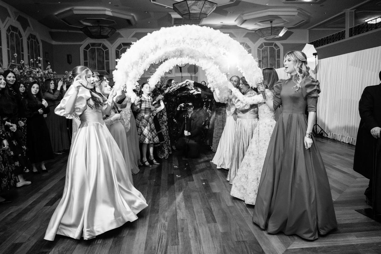 Black-and-white reception dance-floor scene with a bride procession, flowing dresses, and guests cheering on both sides beneath a decorative archway. Wedding photography at The Palace, Brooklyn, NY.