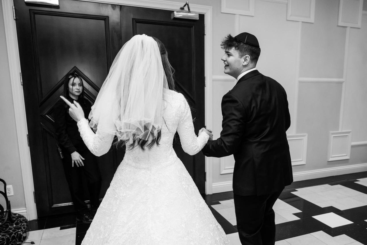 Black-and-white wedding dance moment: bride and groom smiling as they sway hand-in-hand in a hallway near the doors, with a guest watching in the background. Wedding photography at The Palace, Brooklyn, NY.